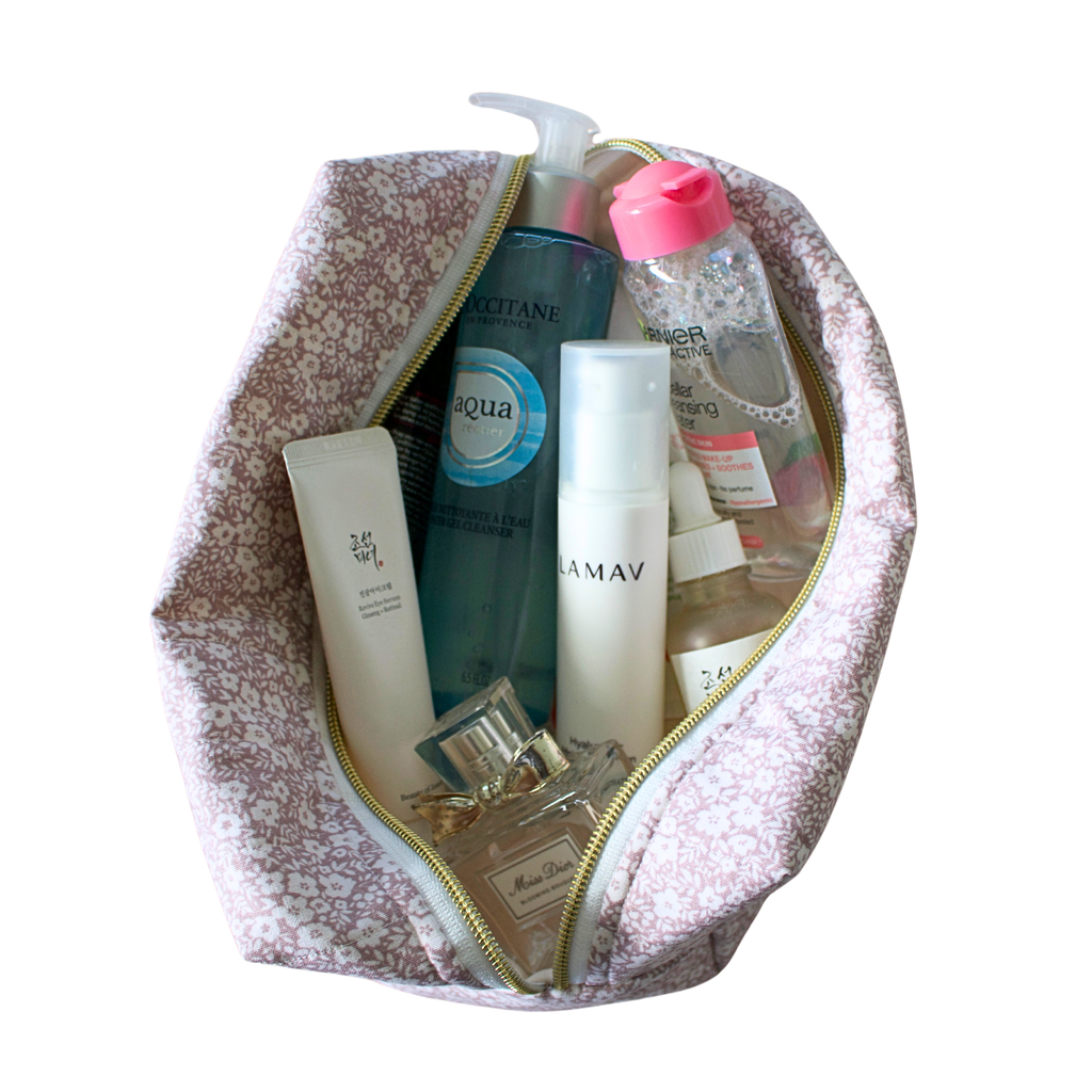 A pink and white flower makeup bag with a gold zipper, shown open and filled with various skincare and cosmetic bottles and tubes. Handmade quilted pouch. handmade cosmetic pouch. Handmade bag.