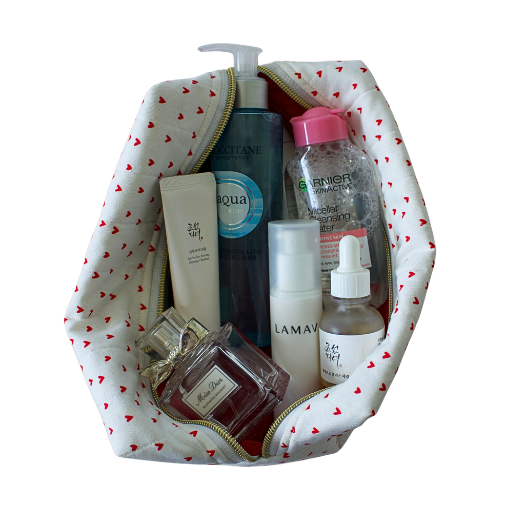 A white cosmetic bag with a red heart pattern, shown open and filled with various skincare and perfume products on a solid white background. handmade pouch. handmade cosmetic pouch. Handmade quilted pouch. Handmade bag