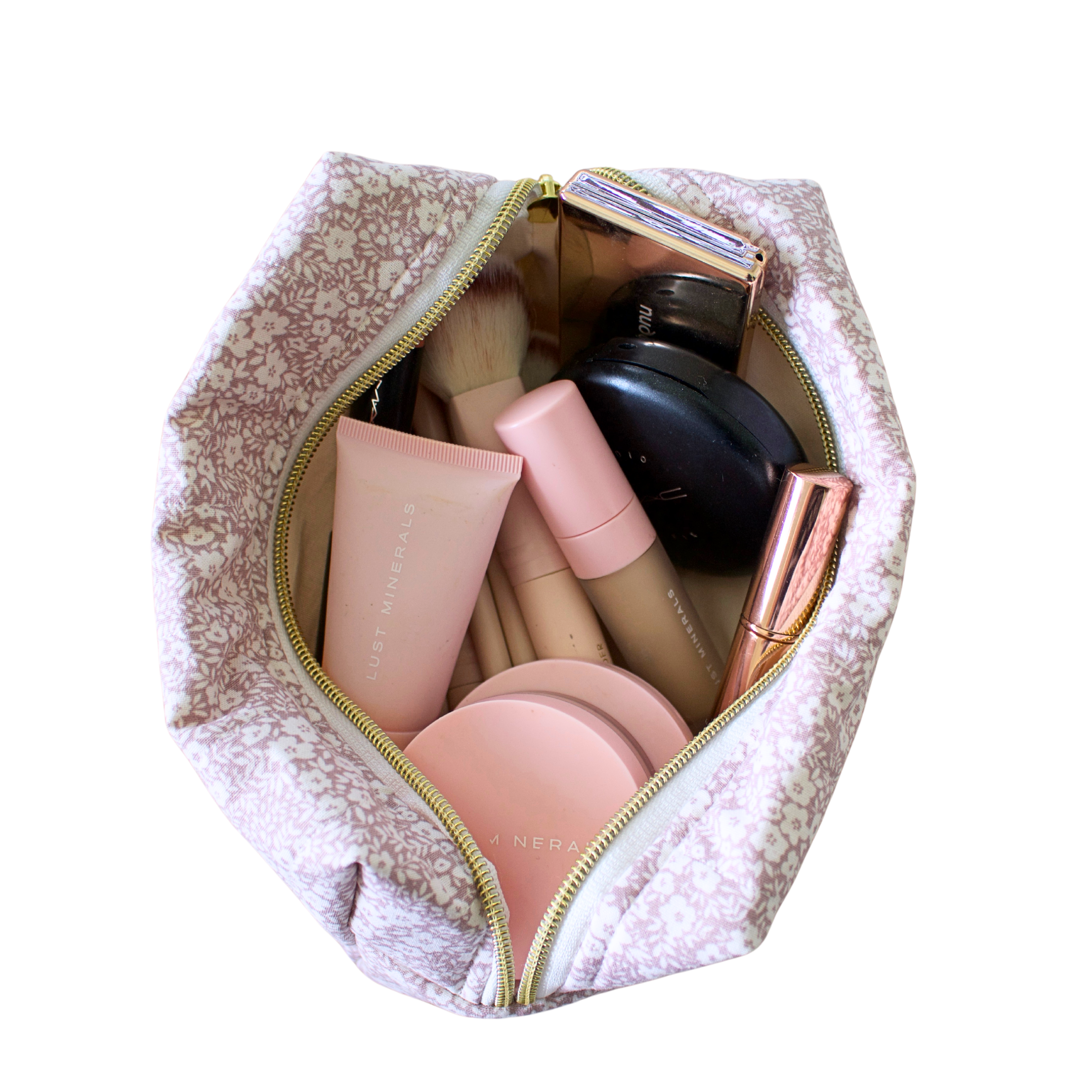 A light pink and white floral makeup bag, shown open from a top-down view, filled with various cosmetic products including brushes, a black compact, and pink tubes, on a solid white background. Handmade makeup bag. Handmade makeup pouch. quilted pouch. Quilted bag