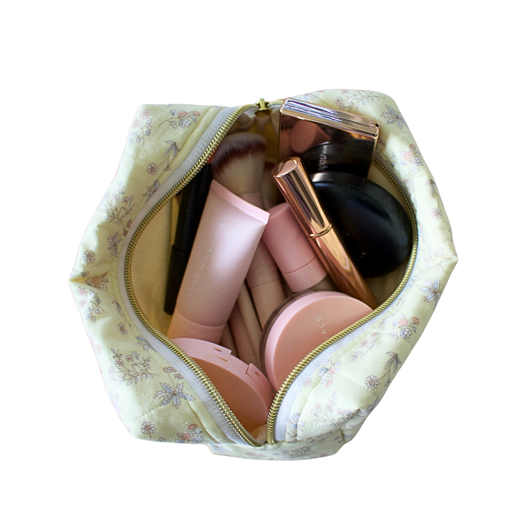 A yellow floral makeup bag filled with various cosmetic products, shown from a top-down view against a solid white background. Handmade quilted Makeup bag. Handmade makeup bag. makeup bag
